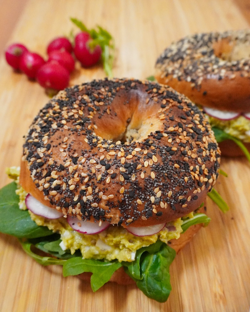avocado bagel sandwich with lettuce and radishes inside in foreground a close up
