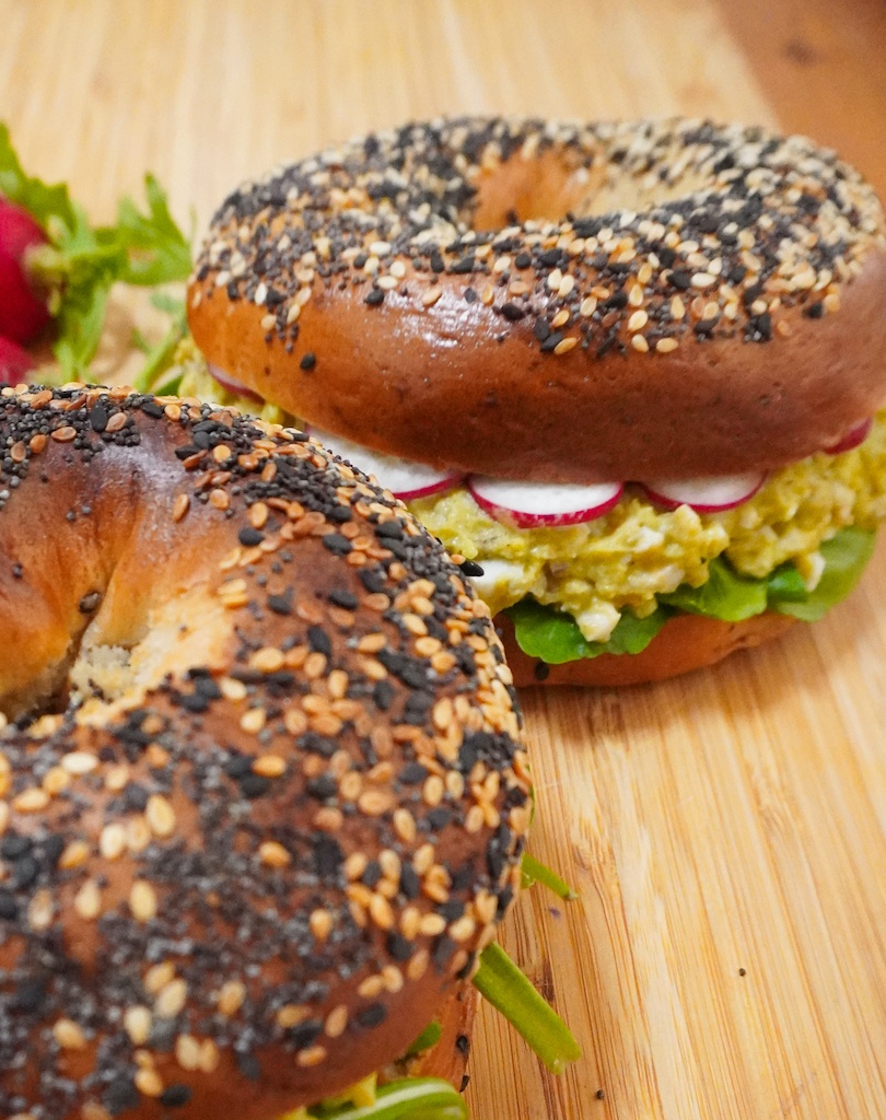 avocado bagel sandwich with lettuce, avocado spread and radishes in in the background with another bagel in foreground