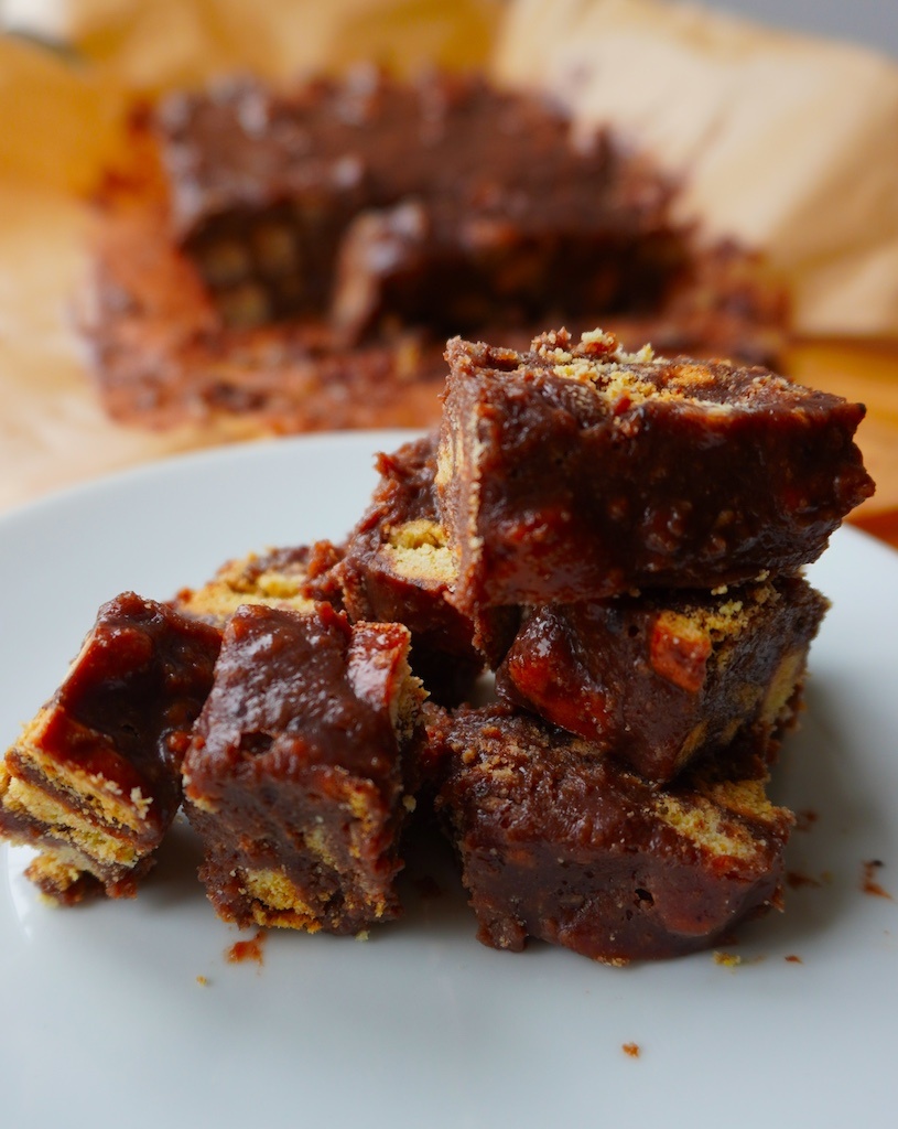 chocolate tiffin cubes in serving plate stacked upon each other more chocolate tiffin in the background