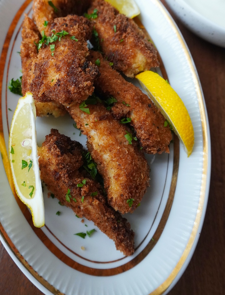 freshly fried fish fingers in oval plate garnished with parsley and lemon wedges close view