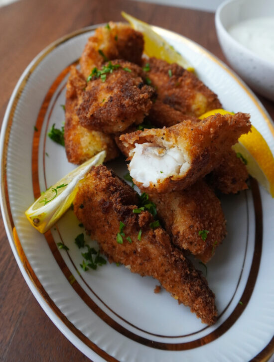 freshly fried fish fingers on oval plate one piece cut open garnished with parsley and lemon wedges