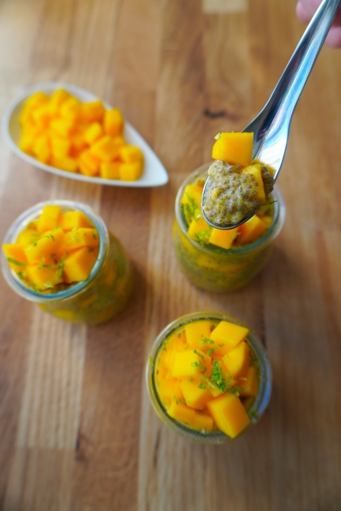 mango chia seeds pudding in glass containers and mango pieces in bowl on wooden worktop with some pudding in spoon