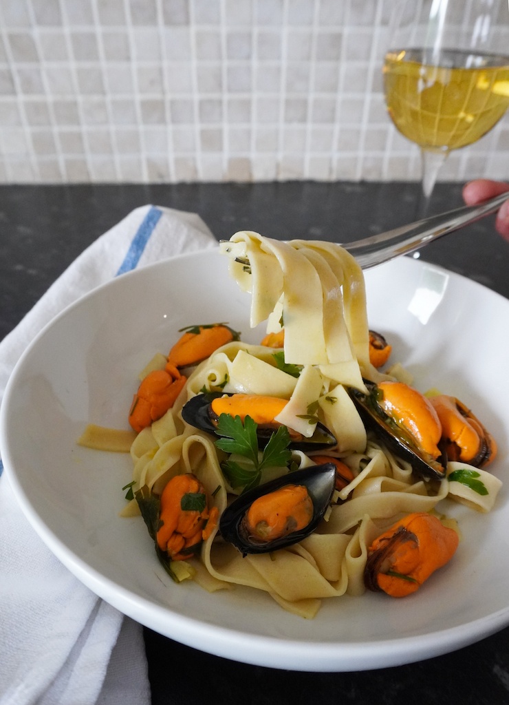 pasta with wine and garlic mussels in pasta bowl hand holding fork with pasta around it glass of wine in background