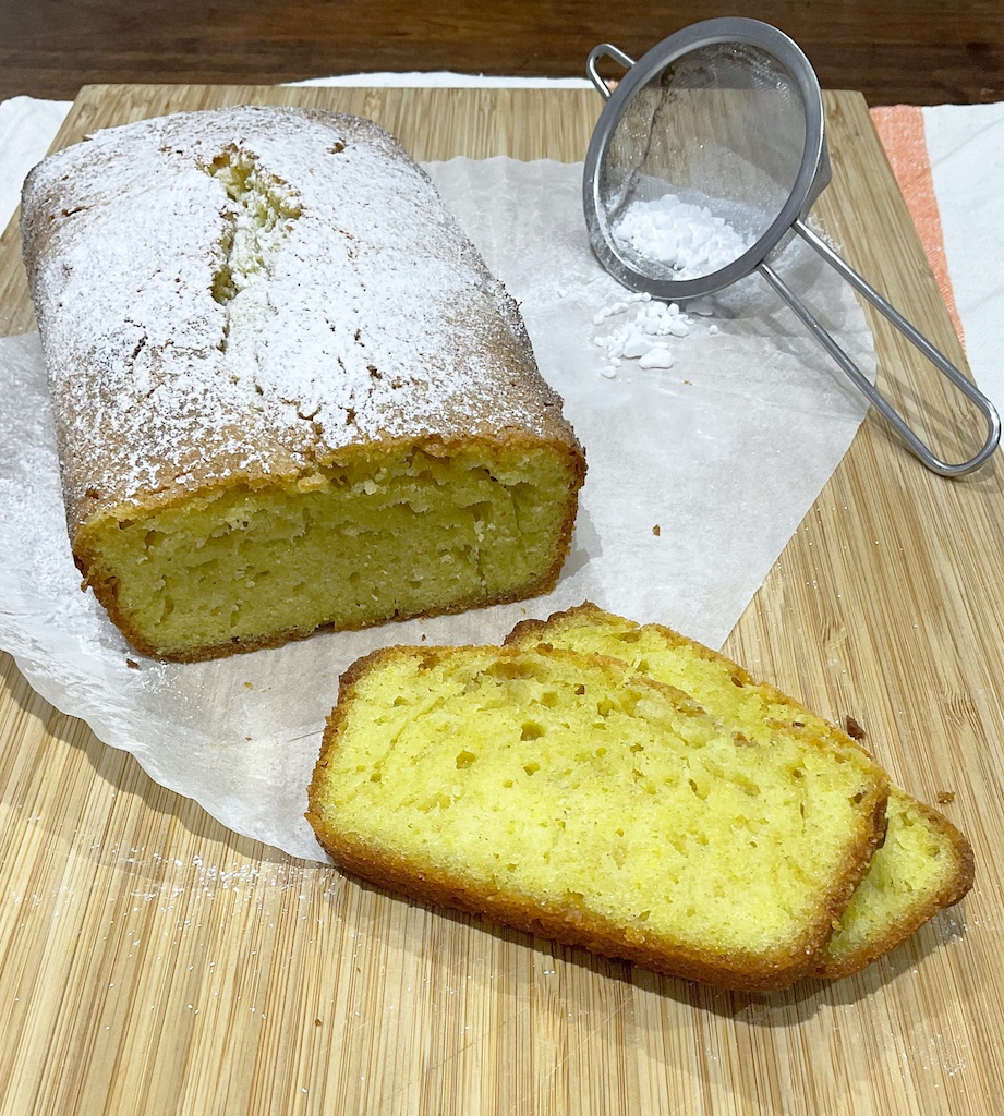 pound cake on the cutting board with two slices and icing sugar in a sieve