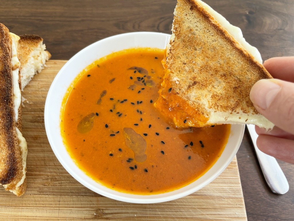 tomato harissa soup with one cheesy toast in the hand dipped in the soup