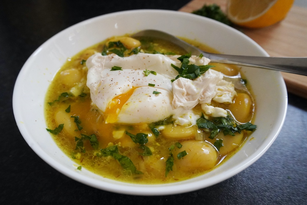 white bean stew with poached egg in serving bowl with a spoon in and lemon in the background